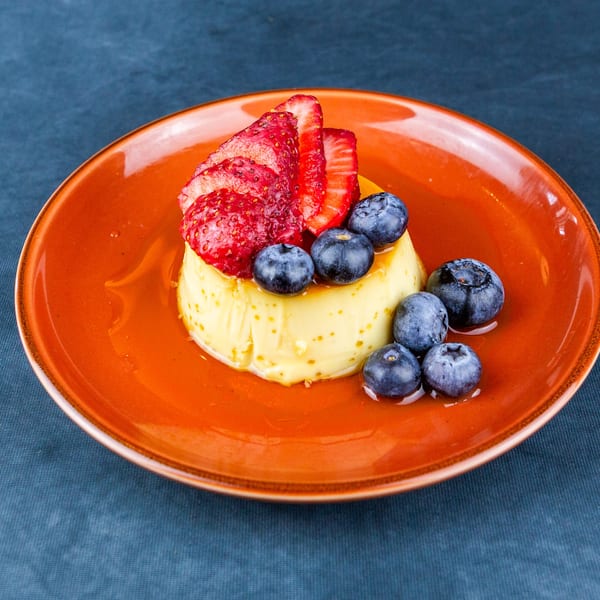 Flan dessert topped with sliced strawberries and blueberries on an orange plate.