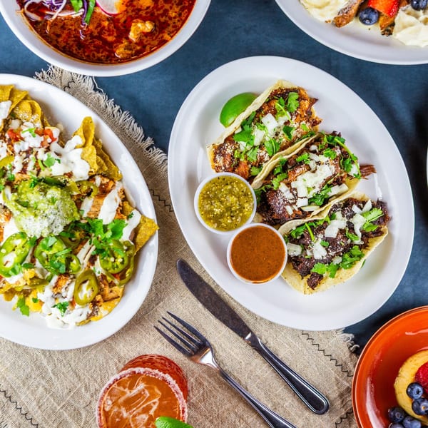 Overhead view of a table laden with Mexican dishes: nachos, tacos, soup, and a drink.