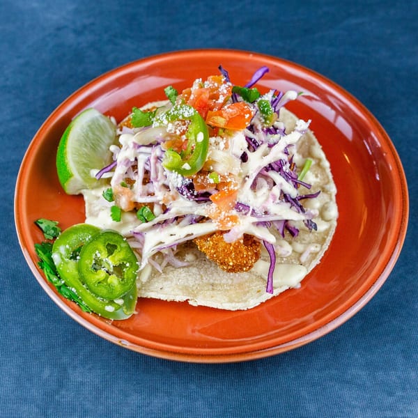 A fish taco on an orange plate, topped with cabbage, salsa, crema, lime, and jalapeños.