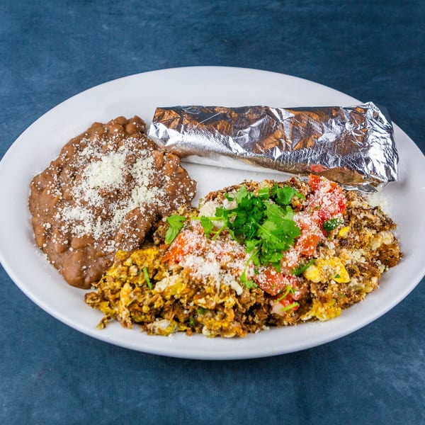 A white plate with refried beans, scrambled eggs with tomatoes and cilantro, and a foil-wrapped burrito.