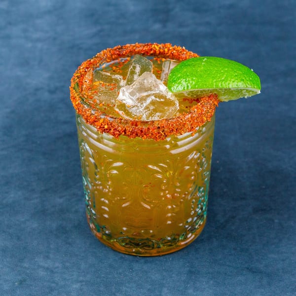 A vibrant yellow drink with ice, a chili-lime rim, and a fresh lime wedge on a blue surface.