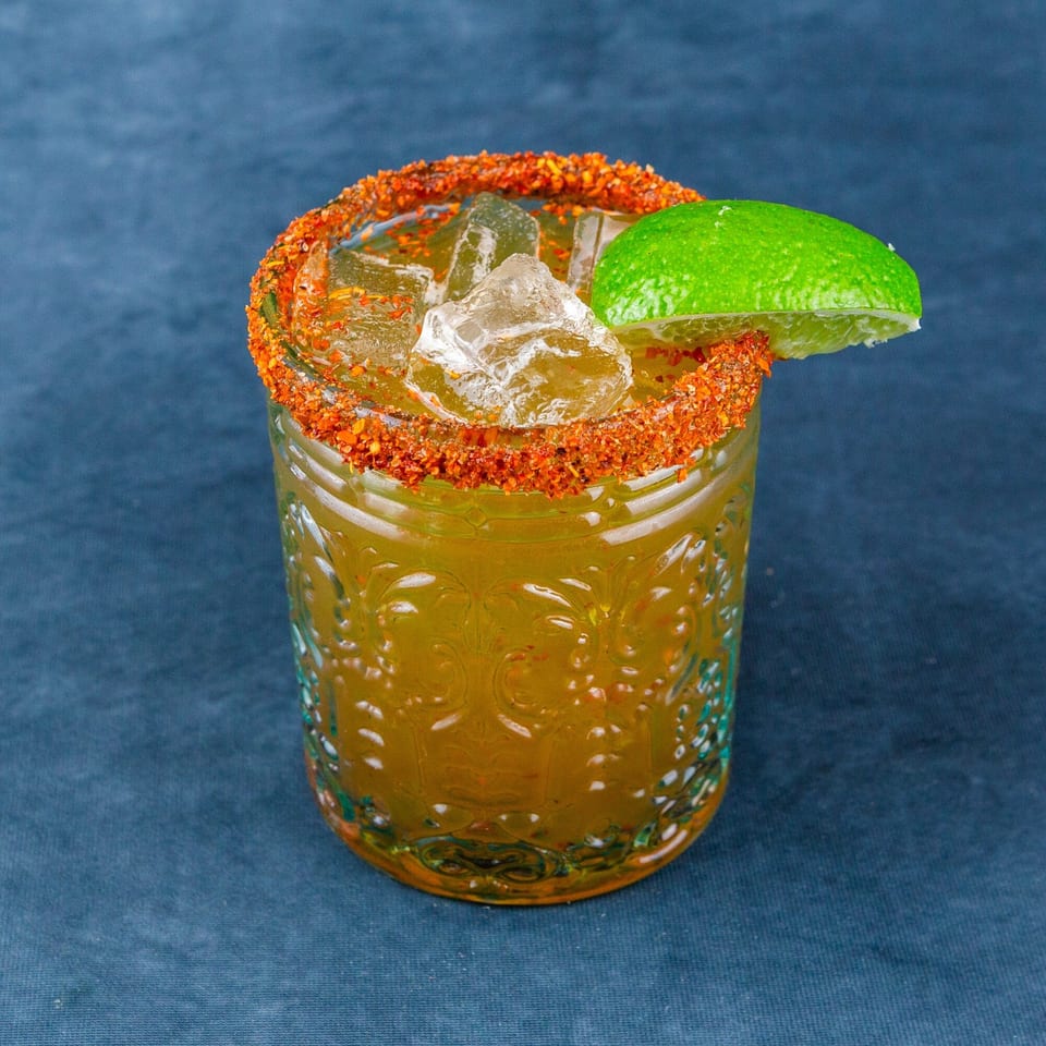 A vibrant yellow drink with ice, a chili-lime rim, and a fresh lime wedge on a blue surface.