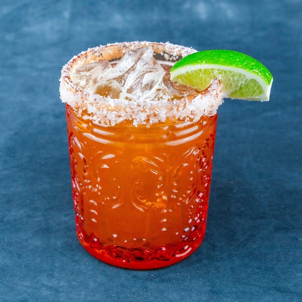 A vibrant orange cocktail in a textured glass, rimmed with salt and garnished with a fresh lime wedge.