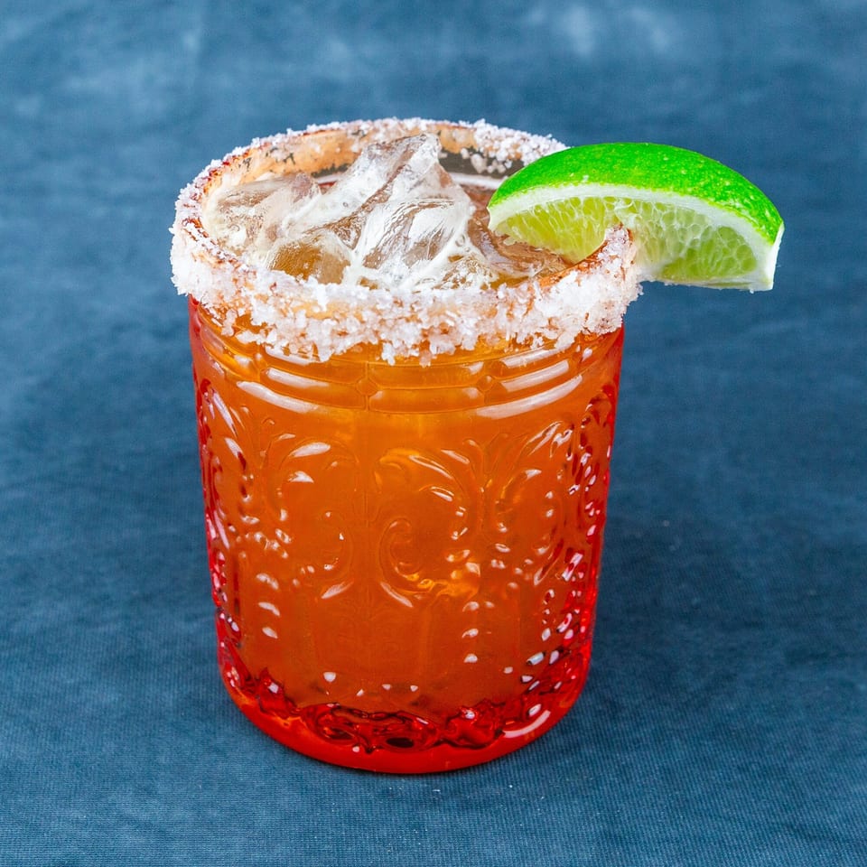 A vibrant orange cocktail in a textured glass, rimmed with salt and garnished with a fresh lime wedge.
