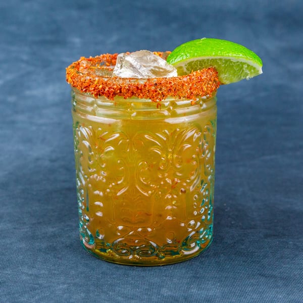 A vibrant yellow drink in an ornate glass, rimmed with chili flakes and garnished with a lime wedge.