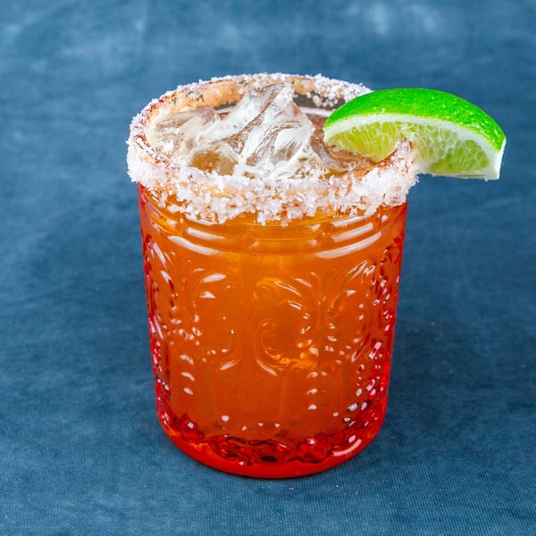 A vibrant orange cocktail in a textured glass, garnished with a salted rim and a fresh lime wedge.